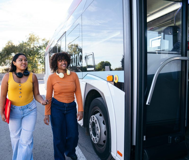 Two young student friends wearing headphones walking towards a public city bus, ready for their daily campus commute