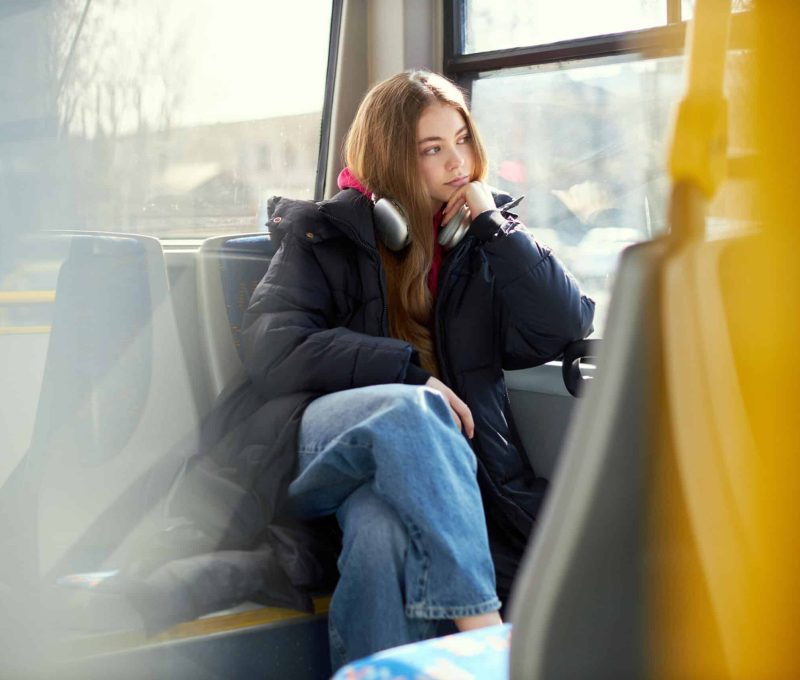 Dreamy looking young woman in headphones sitting in modern tram. Safe transportation. Blurred city streets on background. Concept of public transport, urban lifestyle
